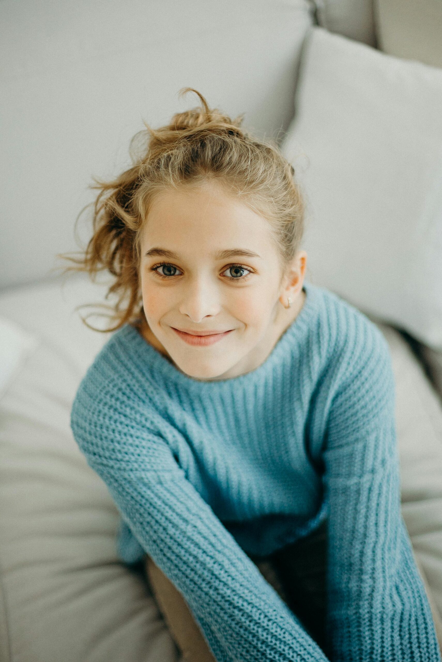 A cheerful young girl with a curly ponytail smiles warmly, wearing a cozy blue knitted sweater.